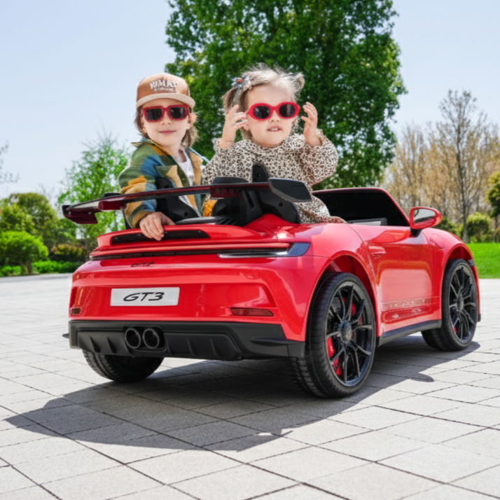 Two children playing in a red PORSCHE 911 toy car on a paved area with trees in the background