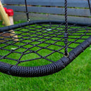 A black rope net swing hanging outdoors over green grass, with a dark wooden fence in the background.