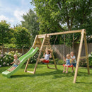 A wooden swing set with a green slide in a backyard, with three children playing on the swings and one sliding down the slide.