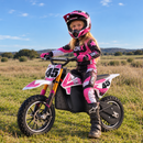 A young girl wearing a pink and black motocross helmet, jersey, gloves, and boots sits on a pink and white electric dirt bike in an open grassy field under a blue sky.