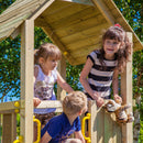 Three children playing on a wooden outdoor playhouse with yellow handles, set against a background of green trees and blue sky.