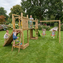 A large wooden children's playground structure in a grassy backyard, with several children playing on the multi-level playset including a slide, climbing walls, rope bridge, and swings.
