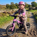 A young child wearing a pink helmet and striped fleece sits on a pink balance bike on a gravel path outdoors, with grassy fields, trees, and a stone wall in the background.