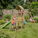 A wooden children's playset in a backyard with several kids playing, including a green slide, climbing ladder, tower, and swing set, surrounded by grass, trees, and a fence.