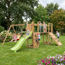 A large wooden children's playground structure in a grassy backyard, featuring a bright green slide, rope swings, climbing wall, rope bridge, and wooden platform. Several children are playing on the equipment under trees and flowering bushes.