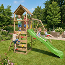 A wooden backyard playset with a green slide, climbing ladder, swing set, and a small house tower, shown outdoors on a grassy lawn with children playing under a blue sky.