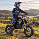 A child wearing a black and blue full-face helmet, matching motocross jersey, gloves, and pants sits on a blue and black electric dirt bike with gold front fork. The child is posed outdoors on a grassy path with a stone wall, lake, and mountains in the background.