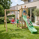 A wooden children's outdoor playset with a green slide, ladder, and hanging swings, shown in a backyard with three children playing on the grass.